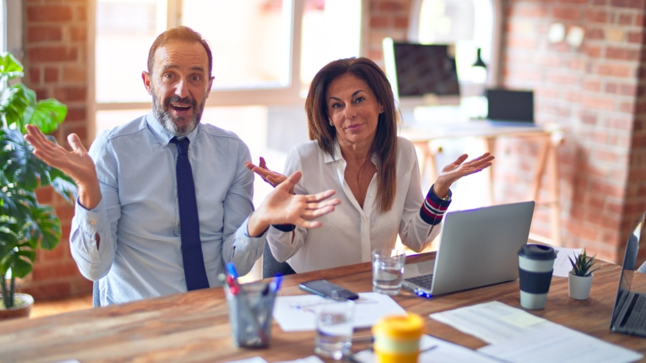 Middle age beautiful business workers working together using laptop at the office clueless and confused expression with arms and hands raised. Doubt concept.